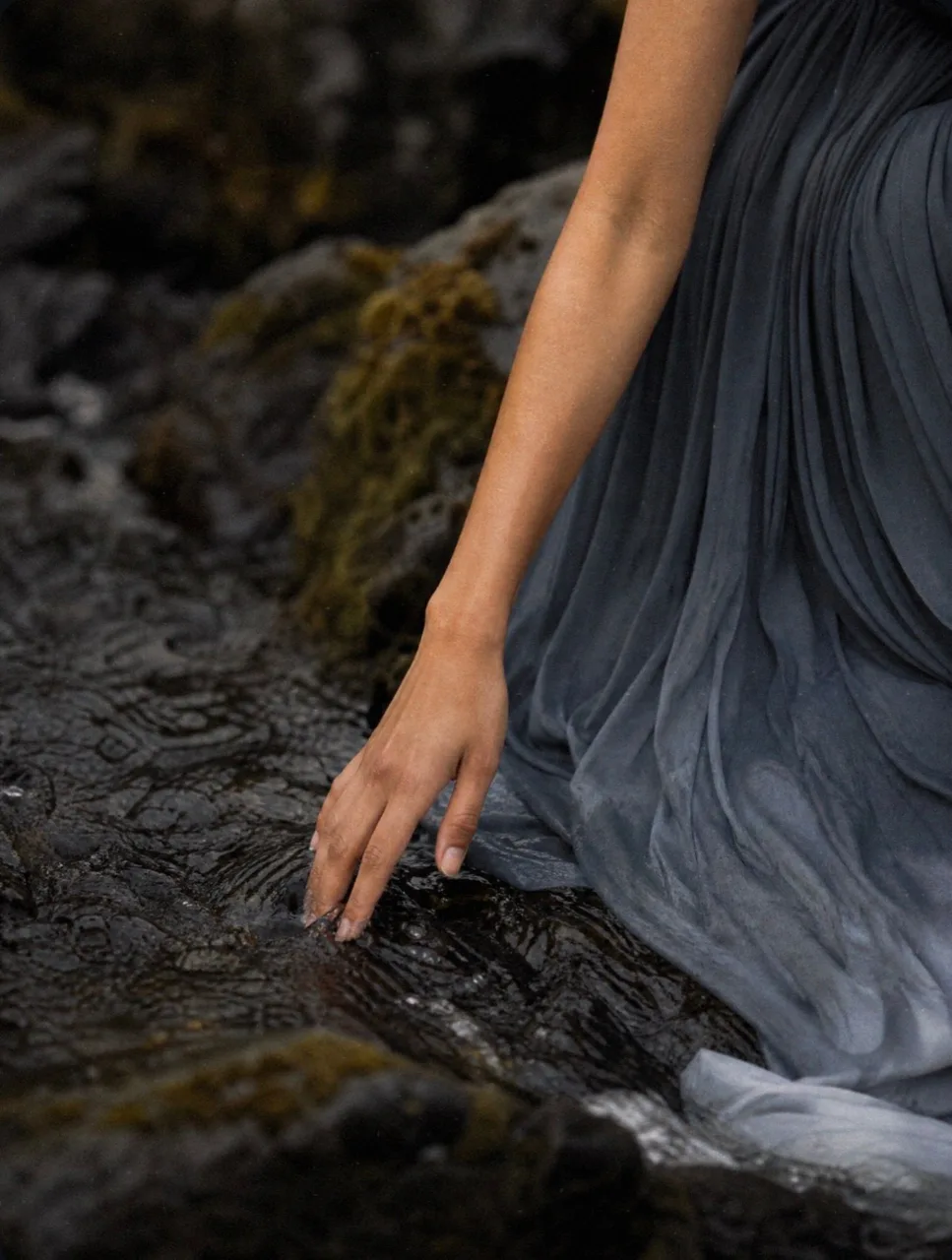 A hand reaching into shallow water beside the hem of a glacial blue gown