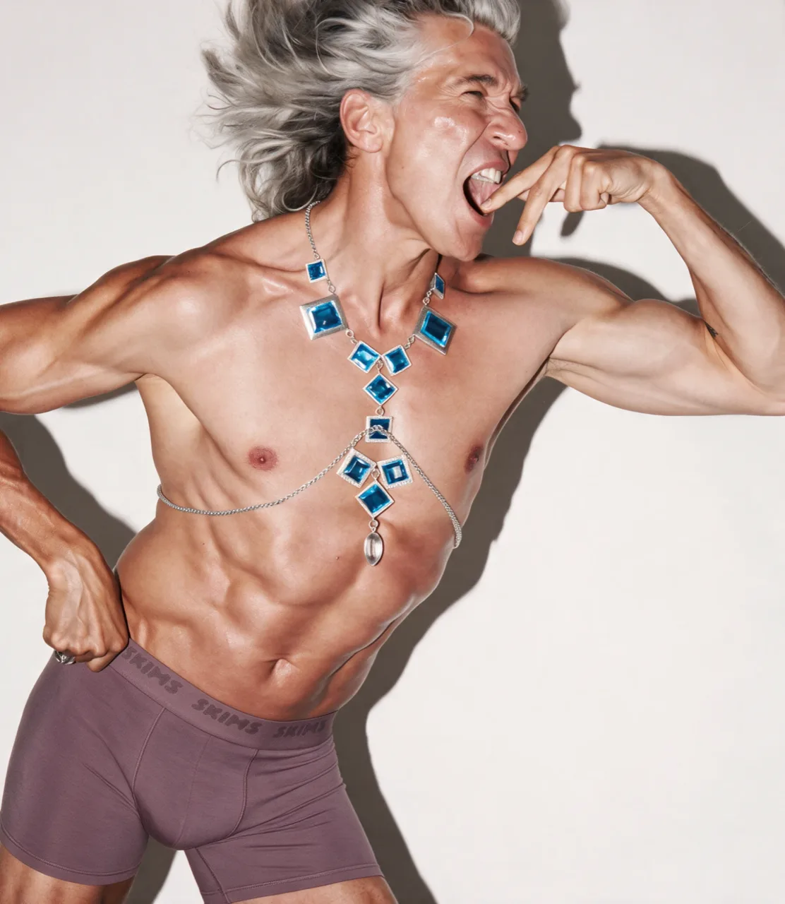 Model with wild grey hair shouting at the lens, a blue gem collar and body chain worn over mauve boxers