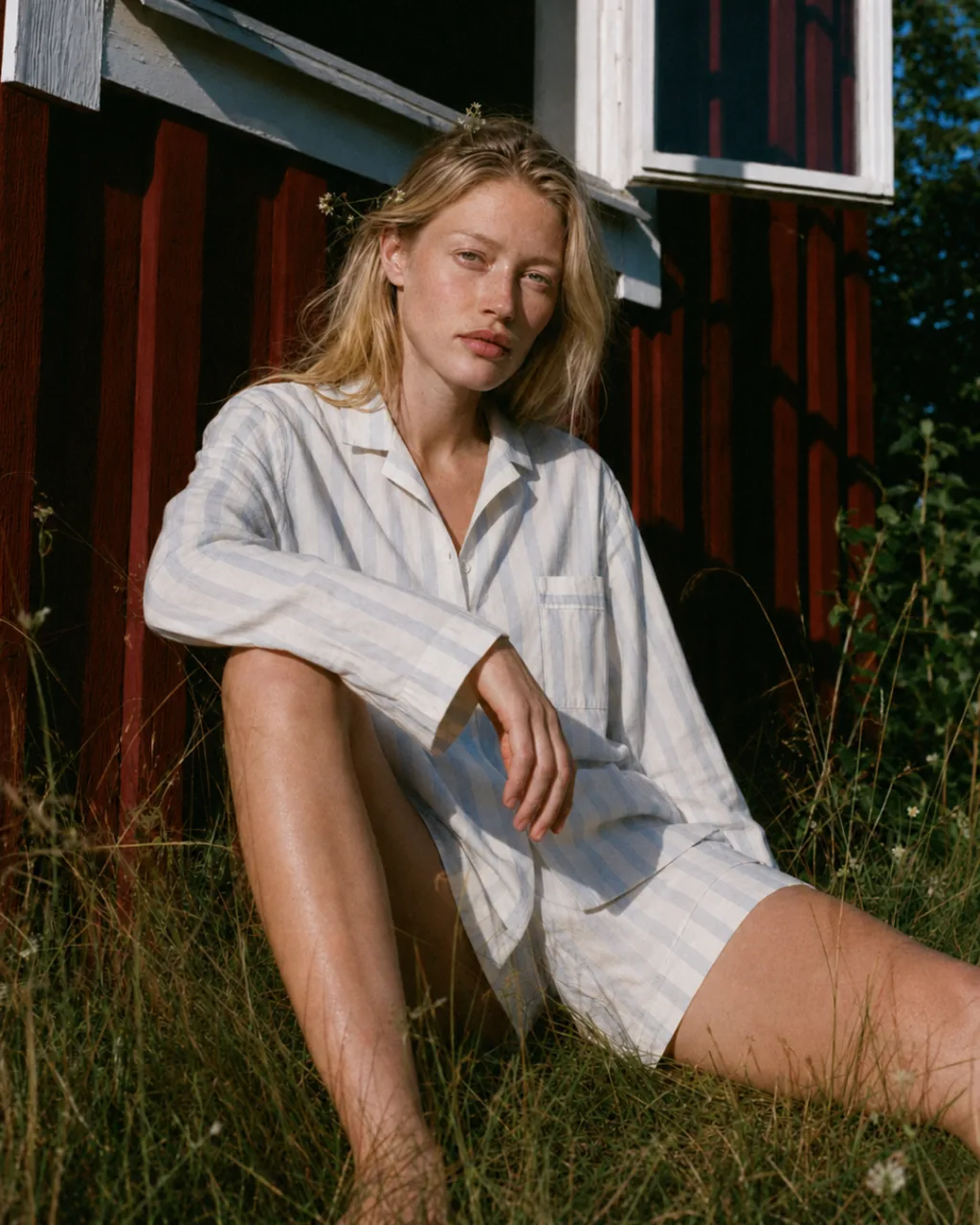 TEKLA summer linens concept campaign, woman in a striped linen shirt sitting in the grass by a red timber cabin