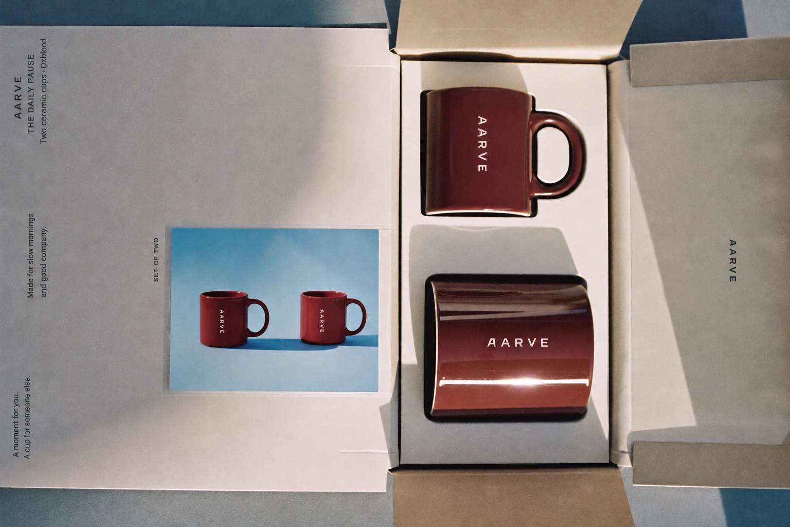 An open gift box holding two oxblood AARVE mugs beside a printed card of the mugs on blue, seen from above, AI concept work