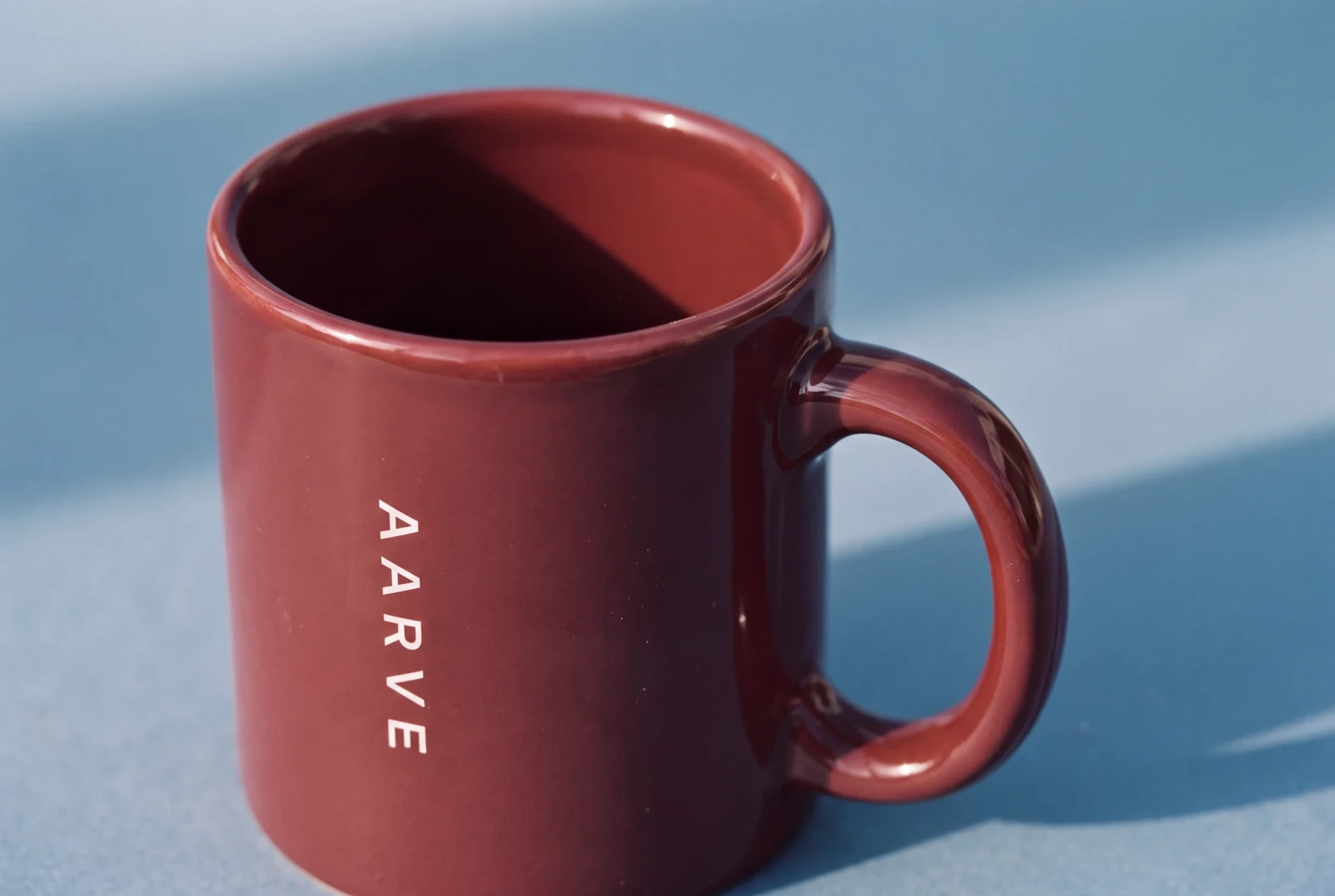 An oxblood ceramic mug printed with AARVE in white, on a pale blue surface in hard sunlight, AI concept work