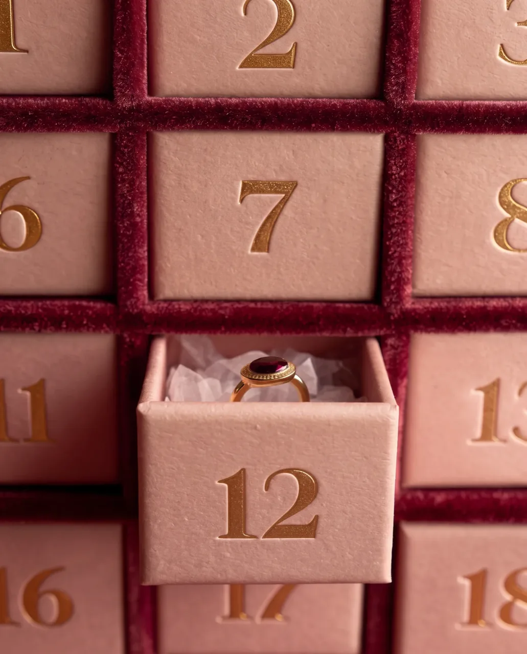An advent calendar with drawer 12 pulled open on a gold ring with a burgundy stone, between burgundy velvet dividers, a Christmas concept frame
