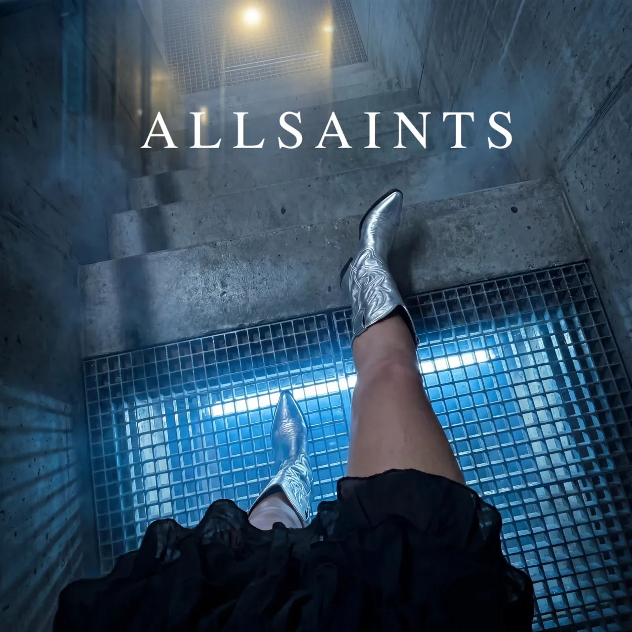 AllSaints key visual: silver cowboy boots on a lit grate in a concrete stairwell, with the AllSaints name, AI concept work
