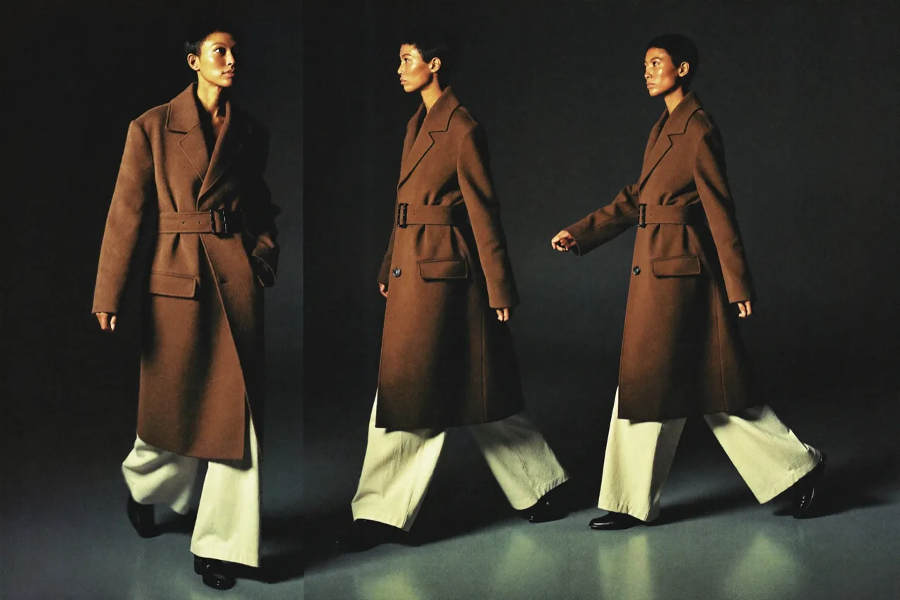 Three frames of the same model in a belted camel wool coat, cream wide-leg trousers and black loafers, walking across a dark studio, once towards the camera and twice in profile