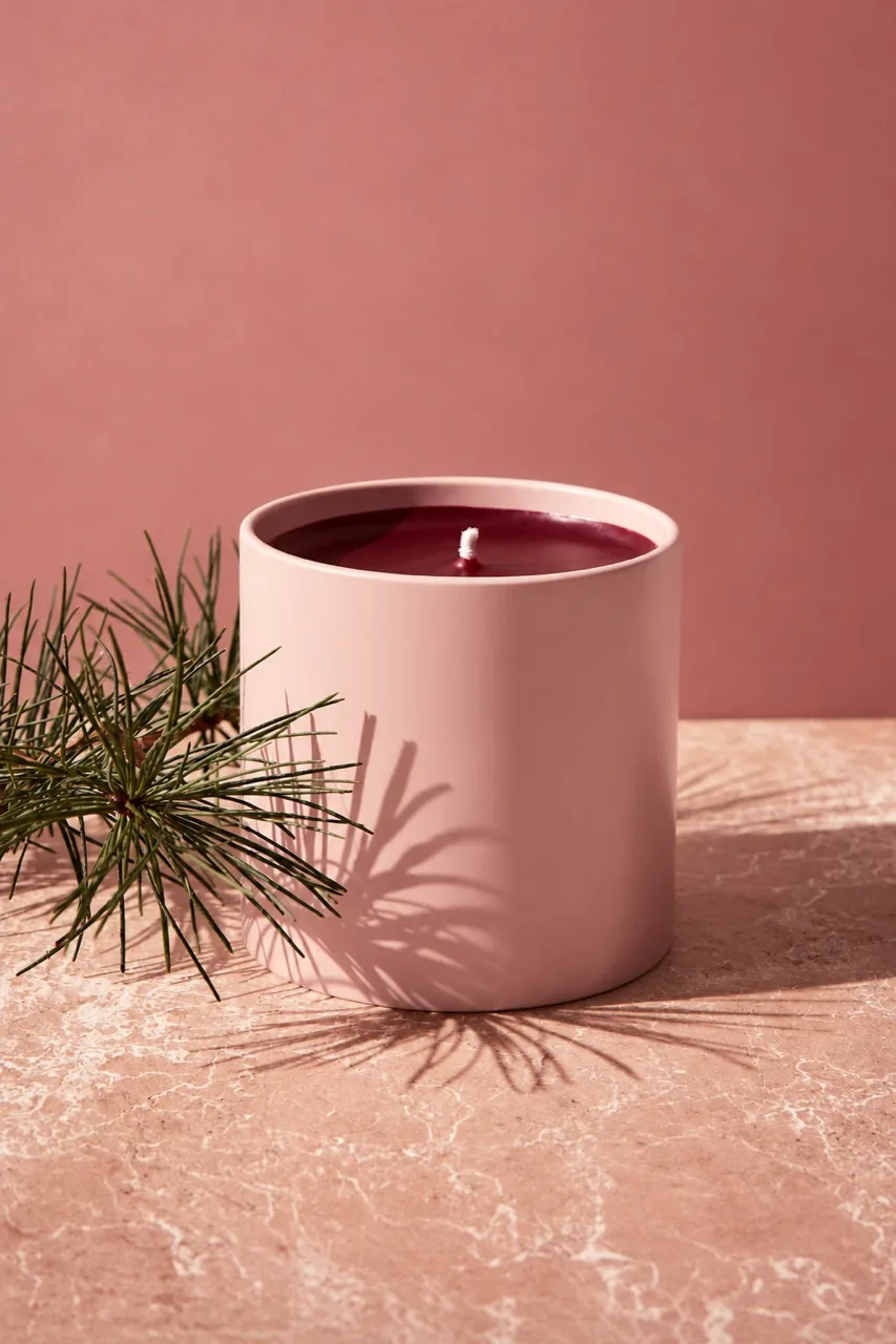 A blush ceramic candle with burgundy wax beside a sprig of pine on pink marble, a minimal Christmas concept frame