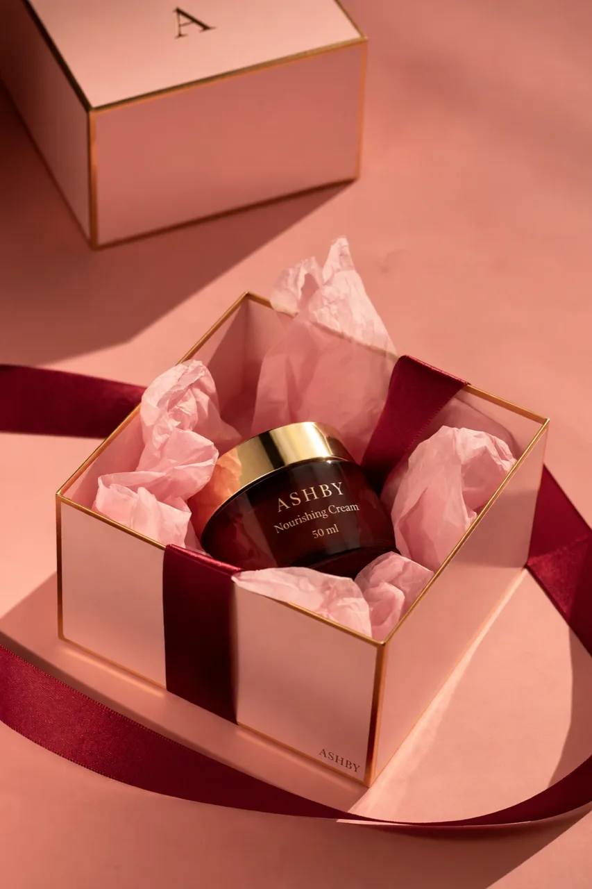 A nourishing cream jar with a gold lid in an open blush gift box with gold edges, on pink tissue with a burgundy satin ribbon, a Christmas concept frame