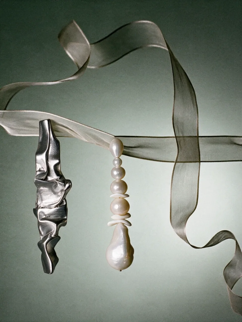 Completedworks concept: a crumpled silver earring and a pearl drop earring hang from a sheer ribbon against a sage green backdrop