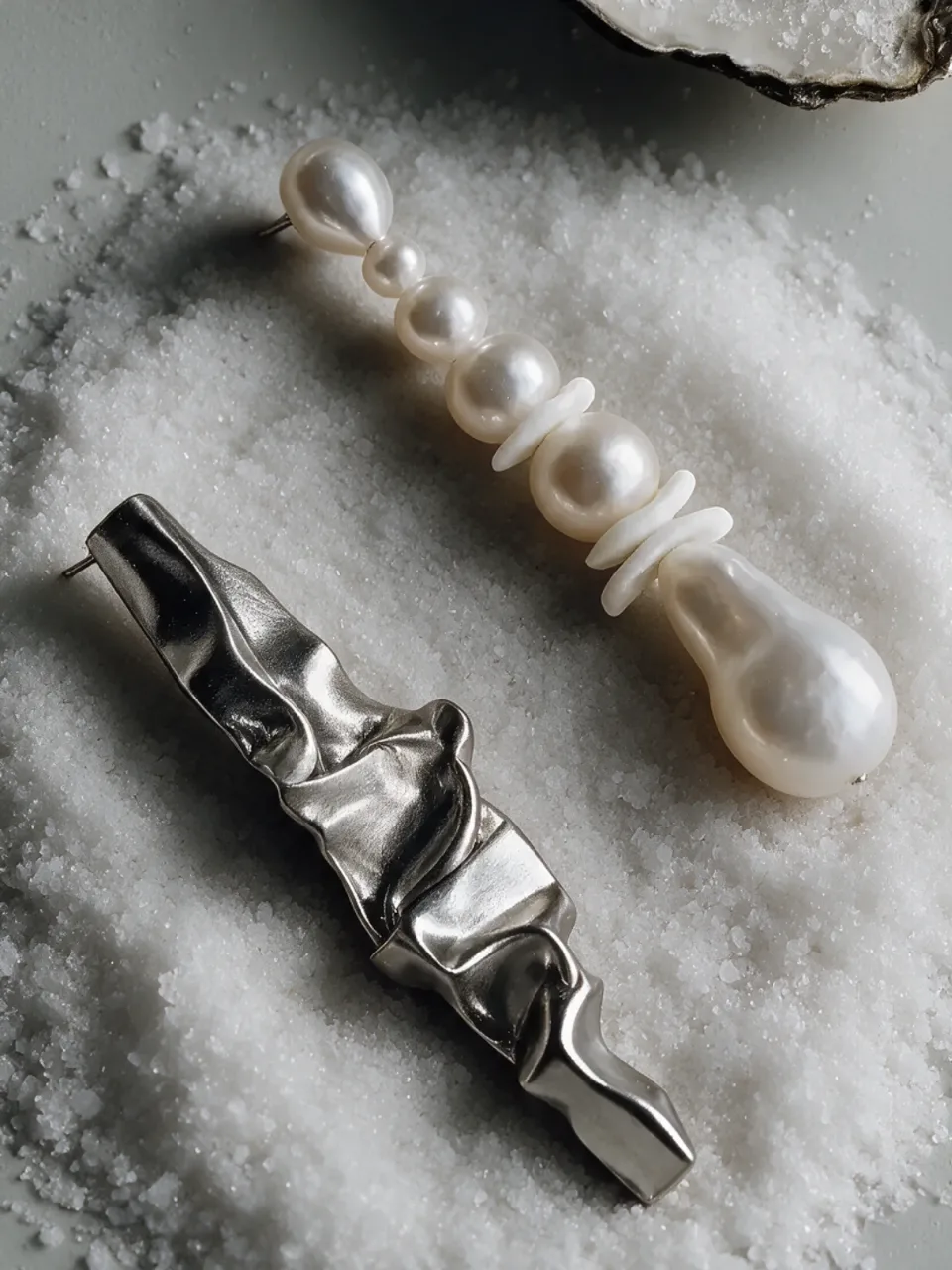 Completedworks concept: a crumpled silver earring and a pearl drop earring on coarse salt beside an oyster shell