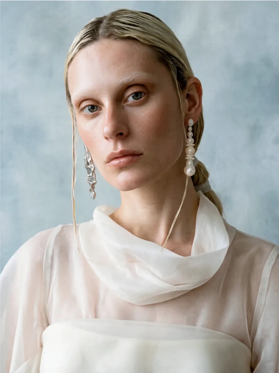 Completedworks concept: a model with slicked-back blonde hair wears a crumpled silver earring and a pearl drop earring, in a sheer white blouse against a pale blue backdrop