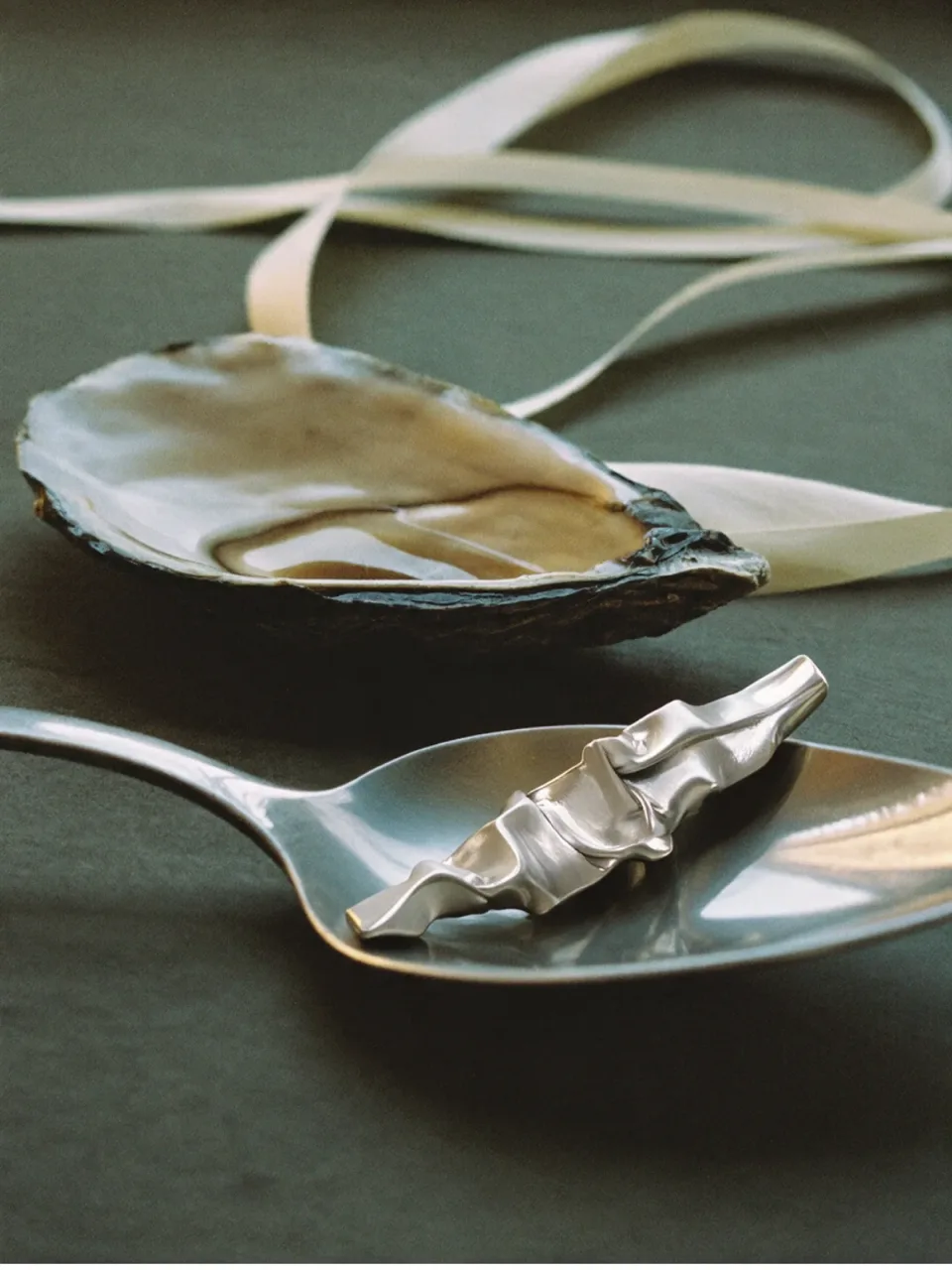 Completedworks concept: a crumpled silver earring rests in a spoon beside an open oyster shell and a cream ribbon