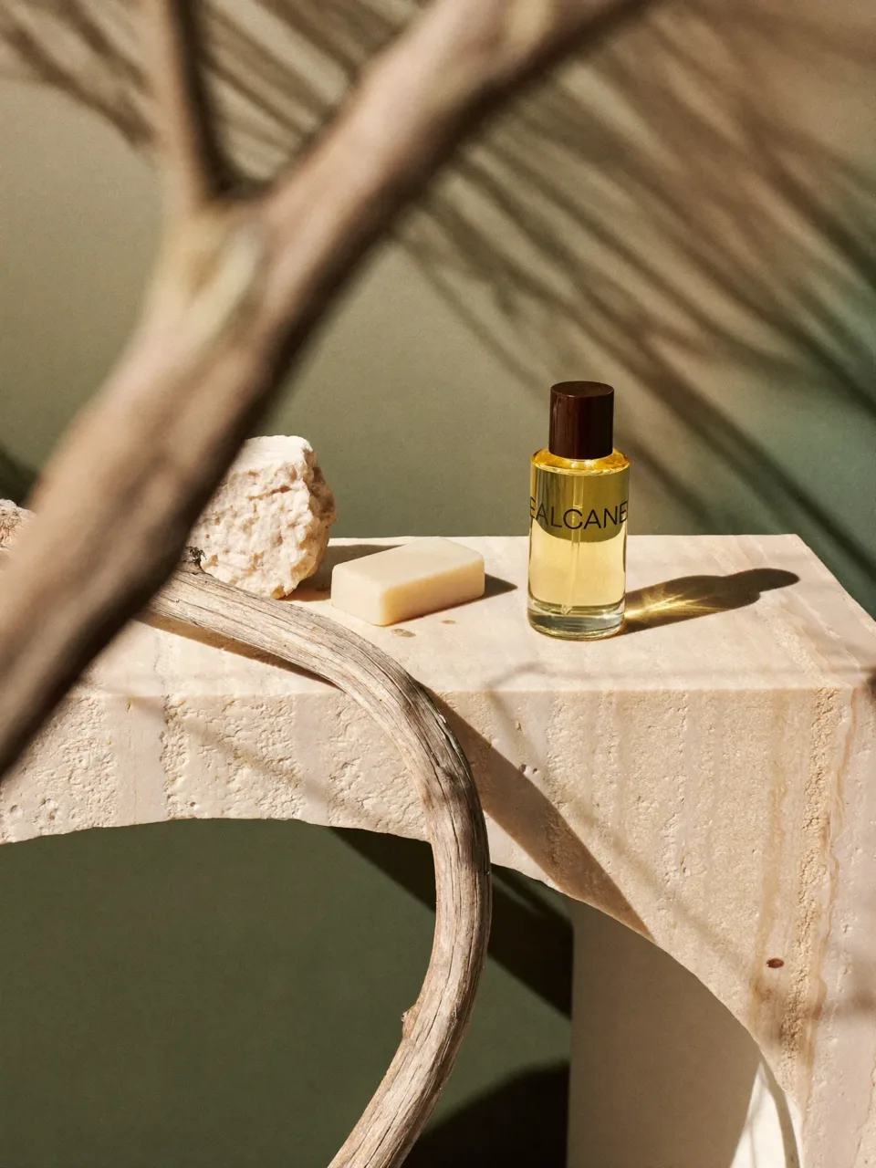 An EALCANE fragrance bottle, a bar of soap and a rough stone on a travertine block, with a driftwood branch and palm shadows on an olive wall