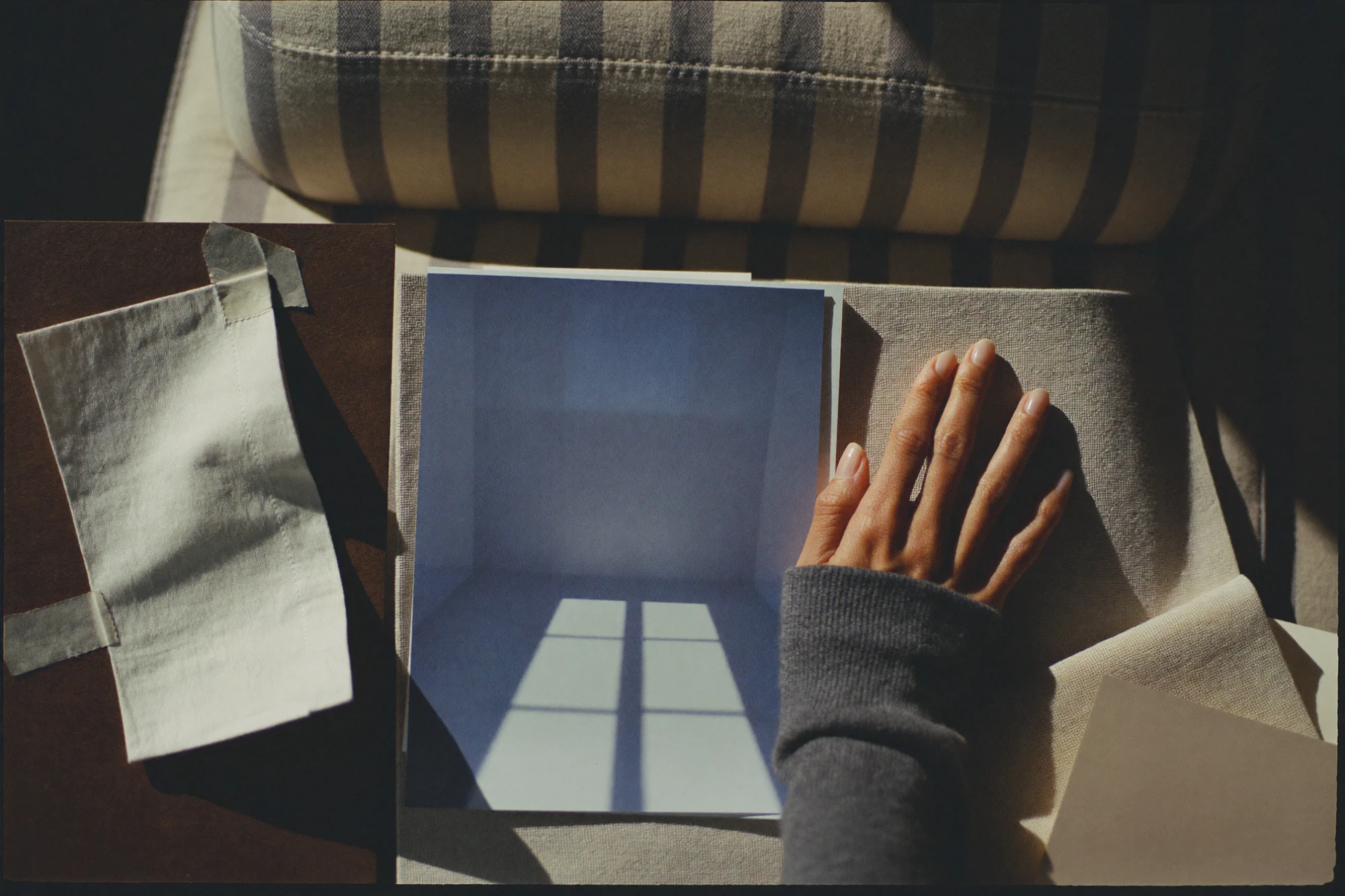 A hand on a blue print of window light, beside a taped linen swatch on a striped armchair, AI concept work