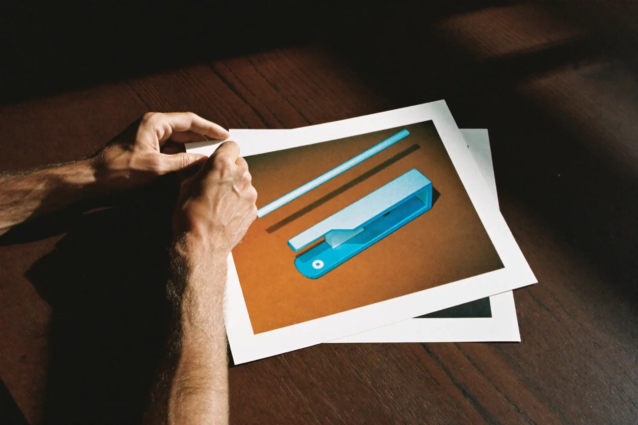 Hands lift a print of a pale blue product in two parts on an orange ground, a second print beneath, AI concept work
