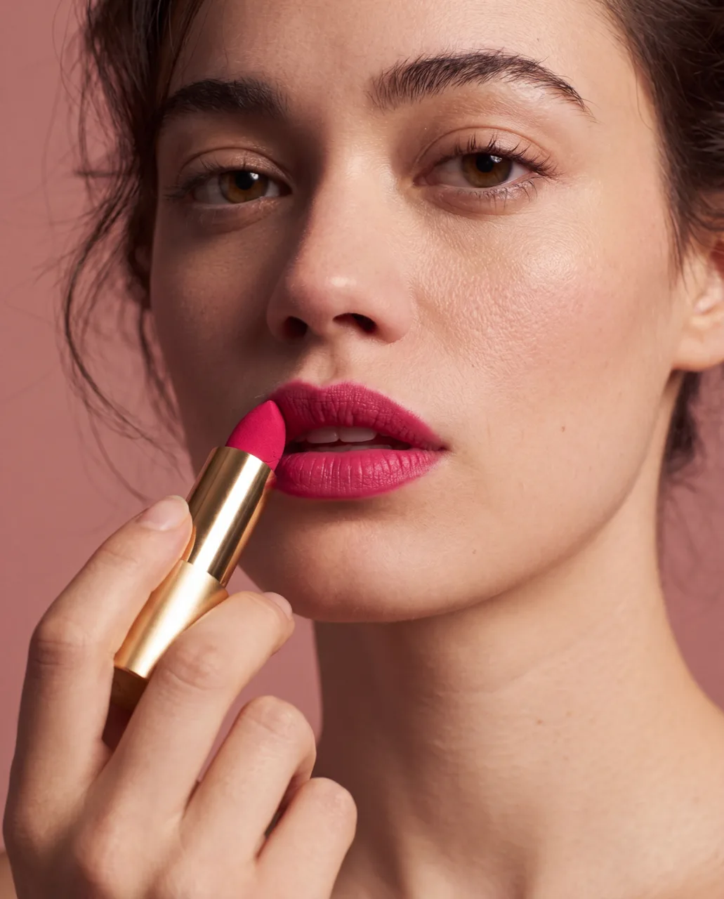 A dark-haired model applies the magenta MUSE lipstick to her lips against a dusty pink backdrop