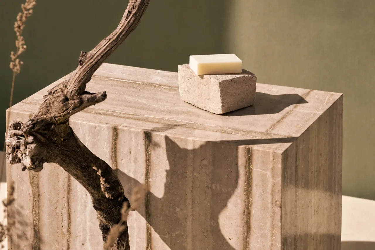 A bar of soap on a rough stone on a veined travertine block, beside a gnarled branch and dried grass