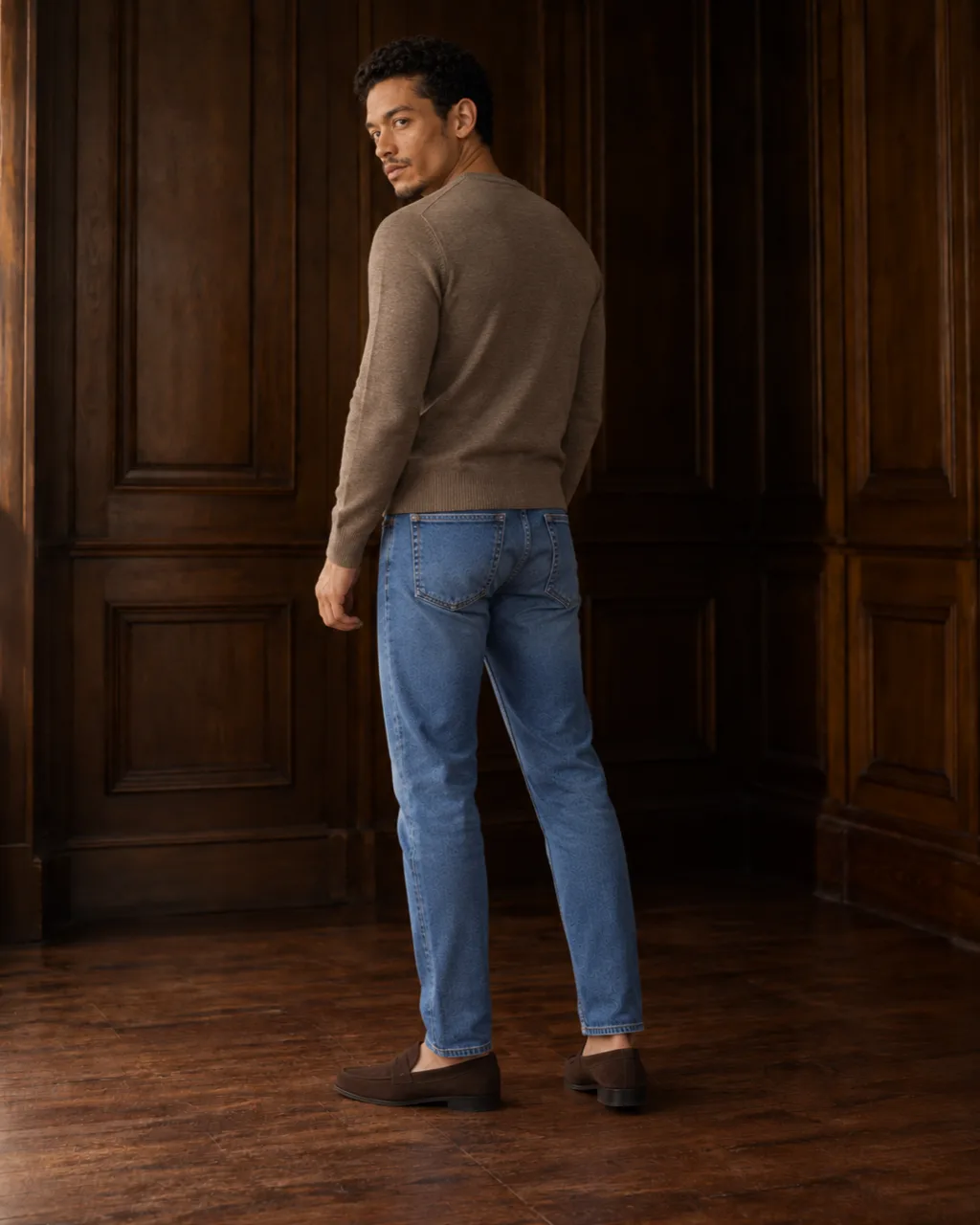 Sunderly look 1, taupe knit and indigo jeans: from behind, three-quarter view, in a wood-panelled room, AI concept work