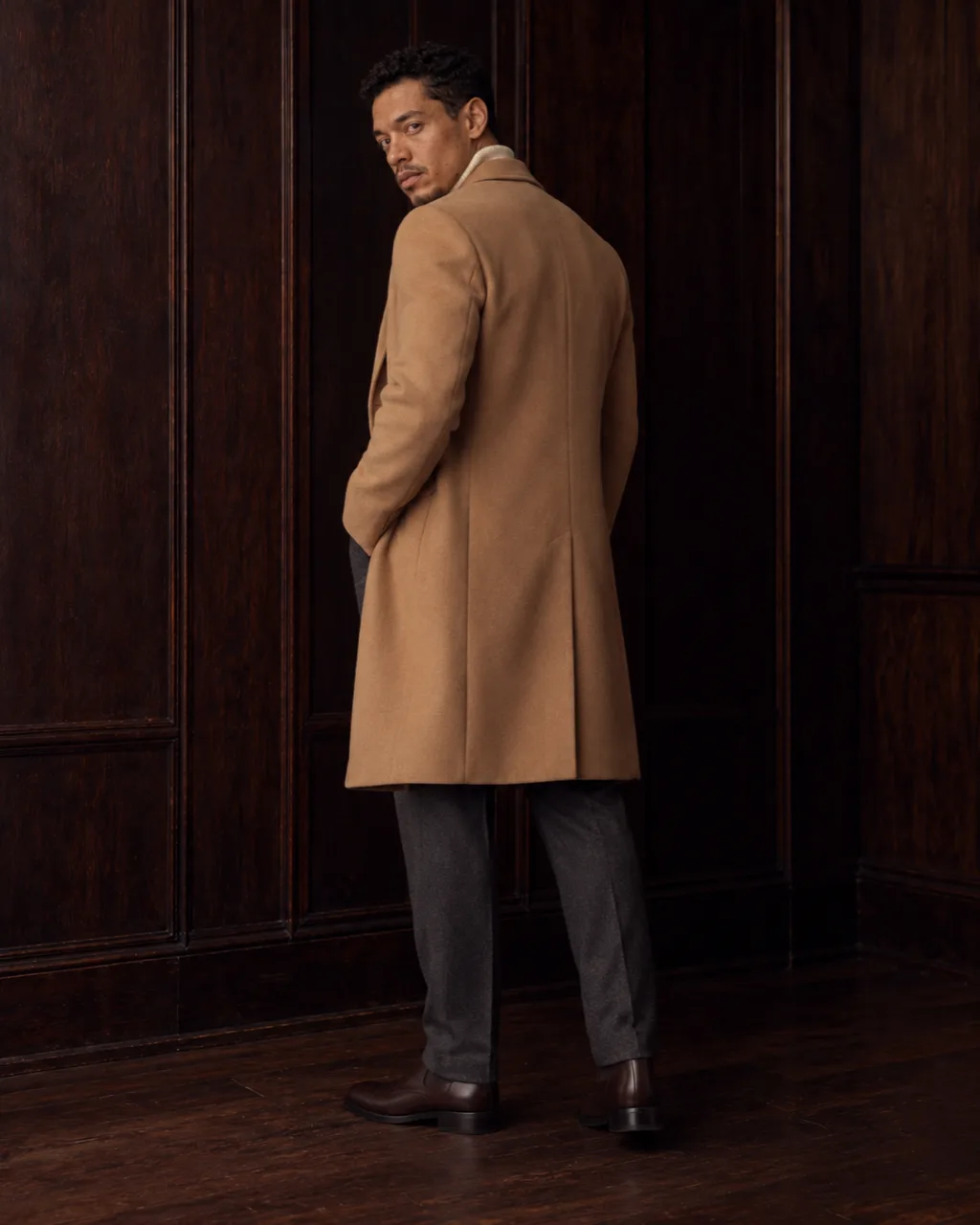 Sunderly look 3, camel coat over a cream roll-neck: from behind, three-quarter view, in a wood-panelled room, AI concept work