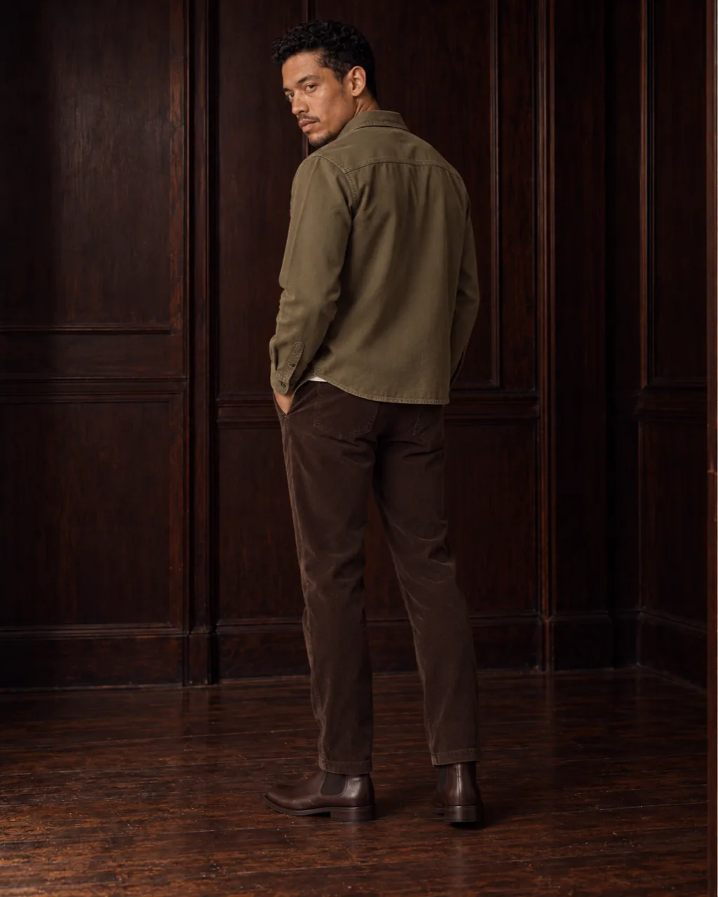 Sunderly look 5, olive overshirt and brown cords: from behind, three-quarter view, in a wood-panelled room, AI concept work
