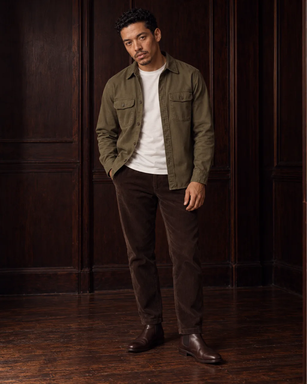 Sunderly look 5, olive overshirt and brown cords: full length from the front in a wood-panelled room, AI concept work