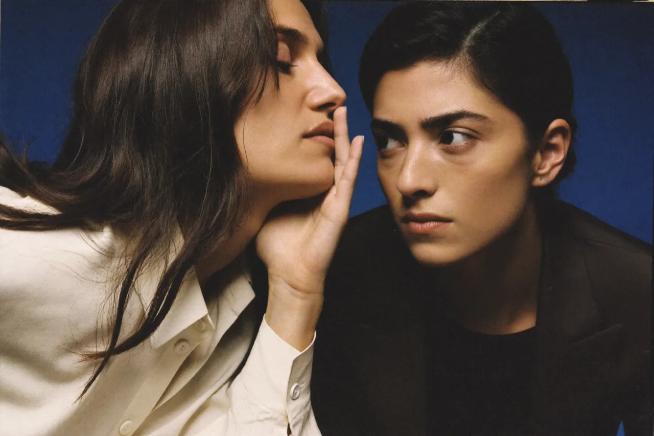 Two models against a deep blue backdrop: one, in a cream shirt, leans in with a hand raised to the lips as if sharing a secret; the other, hair slicked back and in a dark jacket, glances sideways