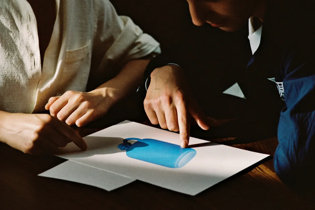 Two people at a wooden table look over a print of a blue bottle, one pointing at the product on the paper, AI concept work